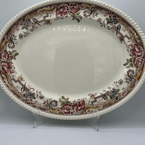 May include: An oval ceramic platter with a cream-colored center and a wide, decorative border. The rim features a rope-like texture and a detailed pattern of pink flowers, brown scrollwork, and small blue birds against a dark brown background.