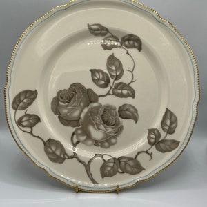May include: A white ceramic dinner plate with a gold rim and a blue trim. The plate features a brown floral design with roses and leaves.