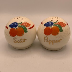Può includere: Un paio di saliere e pepiere vintage in ceramica. Gli shaker rotondi bianchi presentano un design a frutta con accenti arancioni, rossi, blu e verdi. Le parole "Salt" e "Pepper" sono scritte in oro.