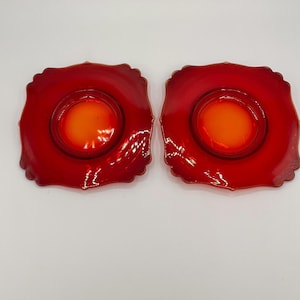 May include: Two red glass saucers with a ruffled edge and a central orange circle. The saucers are symmetrical and appear to be vintage or antique. The glass has a glossy finish, reflecting light.