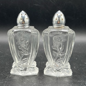 May include: A pair of clear glass salt and pepper shakers. Each shaker has a silver metal top with small holes and a floral design etched into the glass. The shakers sit on a clear, geometric base.