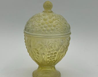 May include: A yellow glass candy dish with a lid. The dish has a pedestal base and is decorated with floral and textured patterns. The lid has a small knob on top. The glass has a slightly frosted appearance.