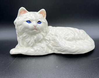 May include: A white ceramic cat figurine in a reclining pose. The cat has blue eyes, pink inner ears, and detailed fur texture. The figurine is set against a dark background.
