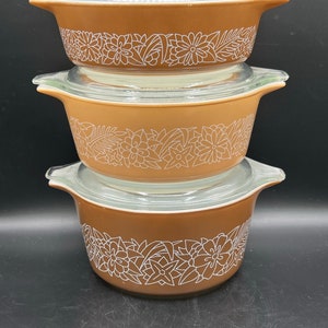 May include: Three brown Pyrex casserole dishes stacked on top of each other, each with a clear glass lid. The dishes have a white floral pattern around the sides. The background is black.