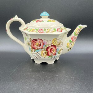 May include: A cream-colored ceramic teapot with a floral design featuring red, yellow, and pink roses. The teapot has a blue knob on the lid and a curved handle. The spout is also decorated with flowers. A charming addition to any tea set.