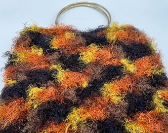 May include: A handmade, textured handbag with a gold-colored metal handle. The bag features a shaggy, multi-colored design in shades of brown, orange, yellow, and black. The bag's unique texture and color palette give it a vintage or bohemian aesthetic.