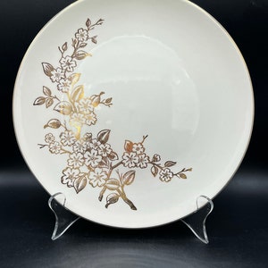 Puede incluir: Un plato de cena de cerámica blanca con un borde dorado y un diseño floral dorado. Las flores son estilizadas y tienen un aspecto delicado y femenino.
