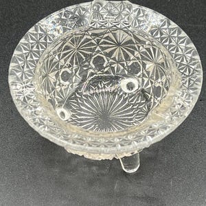 May include: Clear, round glass ashtray with a detailed, geometric pattern. The ashtray has a shallow bowl with a starburst design in the center and a decorative rim. It is supported by three small feet.