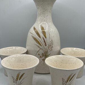 May include: A set of five white ceramic cups and a matching pitcher with a speckled glaze and a floral design featuring wheat stalks.