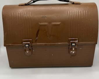 May include: A brown metal lunch box with a black handle and two latches. The lunch box has a raised letter "T" on the front.