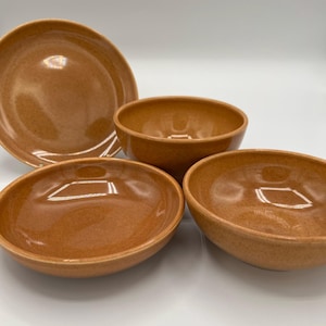 May include: A set of four ceramic dishes in a warm, speckled brown color. The set includes a large plate and three bowls of varying sizes. The dishes have a glossy finish and are arranged on a white surface.