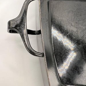 May include: Close-up of a rectangular, dark gray metal baking pan with a curved handle on the left side. The pan has a slightly textured surface and a shiny finish. The handle is also dark gray and has a curved design.