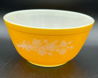 May include: A vintage, round mixing bowl with a bright yellow exterior and a white interior. The bowl features a delicate floral design in white on the outside. This kitchenware item is perfect for mixing ingredients.