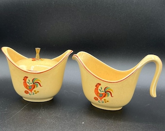 May include: A vintage yellow ceramic sugar bowl with a lid and a creamer. Both pieces feature a rooster design with red, orange, green, and yellow accents. The rims are trimmed with a thin red line.