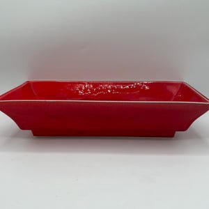 May include: A rectangular, red ceramic serving dish with a white rim. The dish has a slightly flared shape and is set against a white background. The glossy finish reflects light, highlighting its smooth surface.