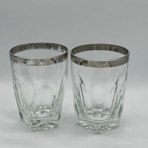 May include: Two clear, faceted drinking glasses with a silver-colored rim. The glasses have a square base and a geometric design. The background is plain white.