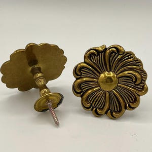 May include: Two antique brass flower-shaped cabinet knobs. One knob is attached to a stem with a screw, while the other is a standalone decorative element. The knobs have detailed petal designs and a warm, golden hue.