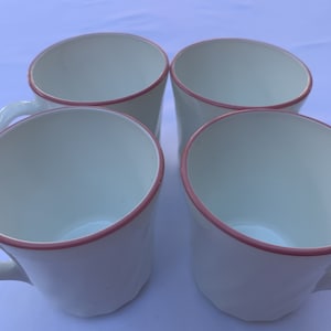 Może przedstawiać: Cztery białe ceramiczne kubki z różowym obramowaniem. Kubki mają unikalny, kątowy kształt i uchwyt. Kubki są ułożone w kwadrat, prezentując czyste linie i prostą elegancję.