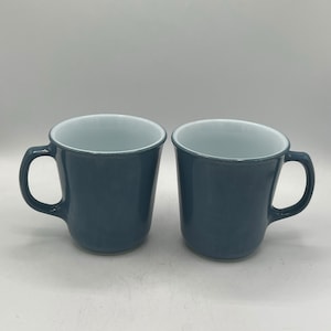 Puede incluir: Dos tazas de cerámica azules con interiores blancos. Las tazas tienen una forma redondeada y un asa en el lado derecho.