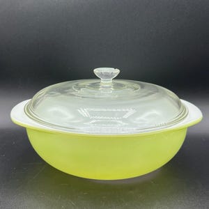 May include: A round, lime green glass casserole dish with a white interior rim and a clear glass lid. The lid features a central knob handle and a subtle, concentric circular pattern etched into the glass surface, providing a clear view inside.