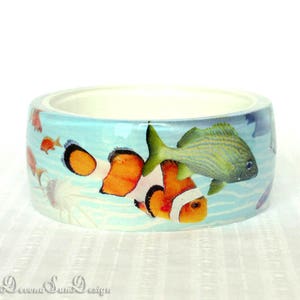 May include: A white ceramic bangle bracelet with a colorful underwater scene featuring orange, white, and black clownfish, a green and blue fish, and other smaller fish. The background is a light blue with white waves.