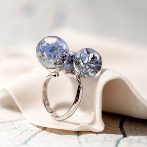 Puede incluir: Un anillo de plata con dos esferas de resina transparente que contienen flores azules de nomeolvides.