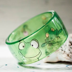 Könnte beinhalten: Ein transparentes grünes Armreif-Armband mit Cartoon-Frosch-Illustrationen. Das Armband zeigt zwei lächelnde Frösche mit großen Augen und einem verspielten Design. Das Armband besteht aus glänzendem Material.