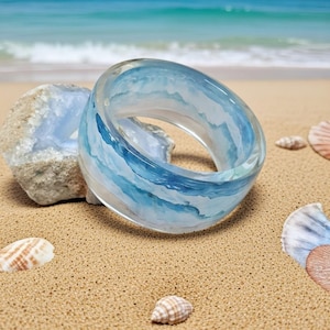 Op de afbeelding: Een heldere harsring met een blauw en wit golfontwerp. De ring ligt op een zandige ondergrond met schelpen en een blauwe steen. De oceaan is zichtbaar op de achtergrond.
