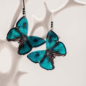 Op de afbeelding: Een paar turquoise blauwe vlinder oorbellen met zilveren haakjes en groene kristallen accenten.
