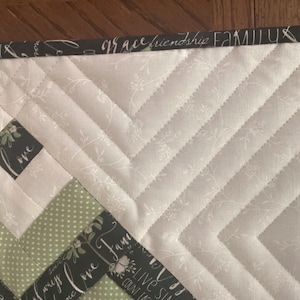 Pode incluir: Uma pe&ccedil;a de tecido acolchoado com um padr&atilde;o de chevron em creme com estampas florais brancas, bolinhas verde-s&aacute;lvia e uma borda cinza-carv&atilde;o. A borda est&aacute; impressa com as palavras family, grace, friendship, love e live simply.