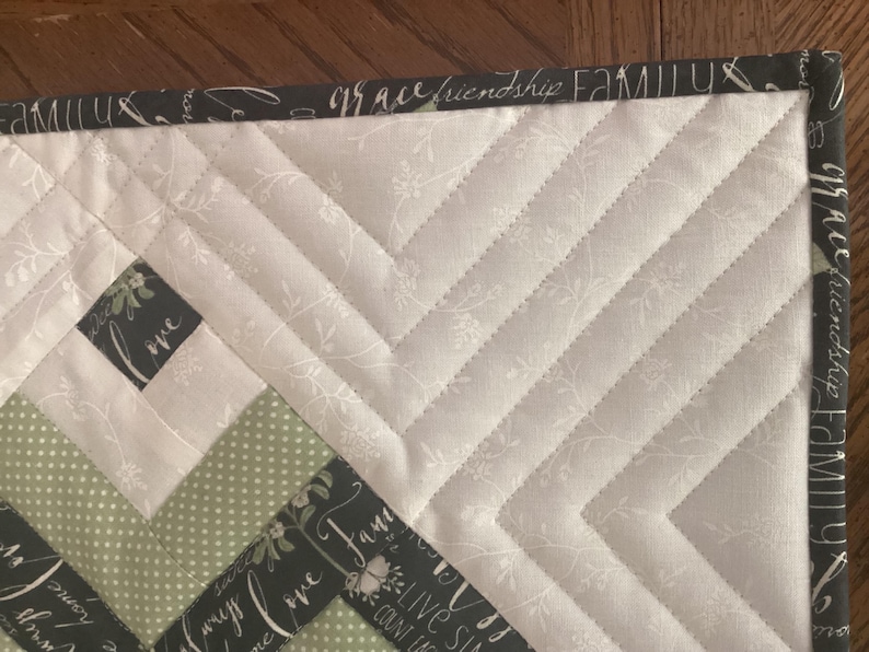 Pode incluir: Uma pe&ccedil;a de tecido acolchoado com um padr&atilde;o de chevron em creme com estampas florais brancas, bolinhas verde-s&aacute;lvia e uma borda cinza-carv&atilde;o. A borda est&aacute; impressa com as palavras family, grace, friendship, love e live simply.