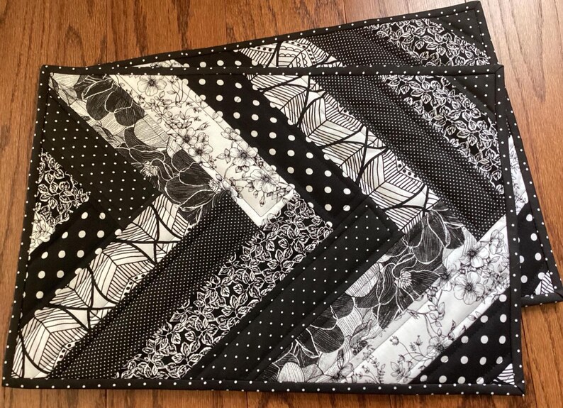 May include: Two black and white quilted placemats with a geometric pattern of black and white floral designs, leaves, and polka dots.