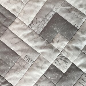 May include: Close-up of a quilted fabric with a diagonal pattern. The design features alternating light gray and white rectangular blocks. Some blocks have floral and script patterns. The stitching is visible, creating a textured surface.