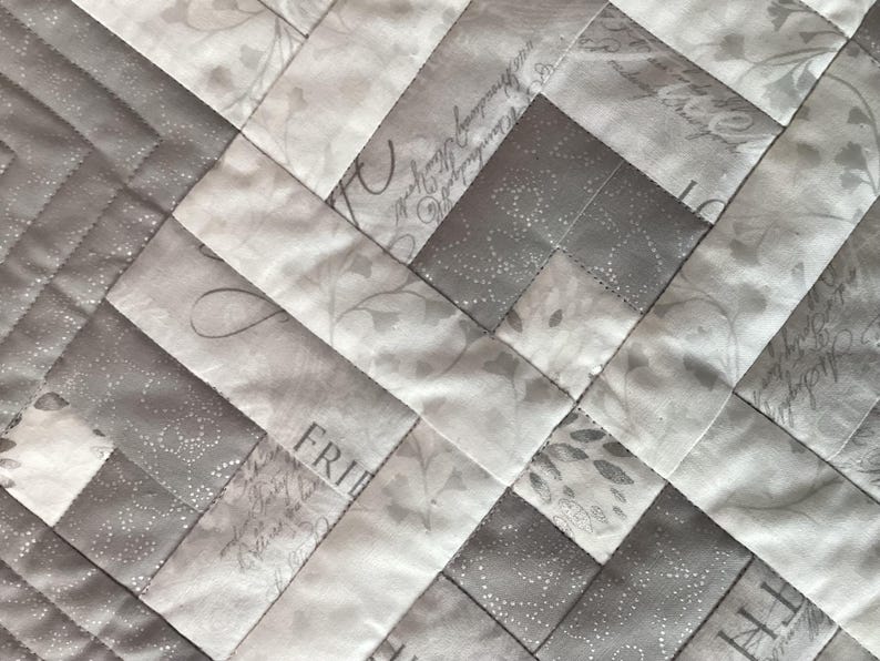 May include: Close-up of a quilted fabric with a diagonal pattern. The design features alternating light gray and white rectangular blocks. Some blocks have floral and script patterns. The stitching is visible, creating a textured surface.