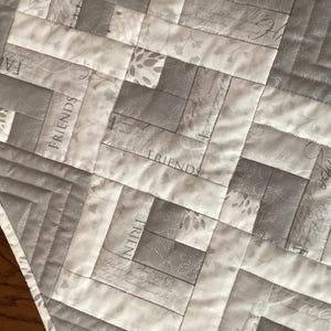 May include: A quilted table runner in shades of gray and white, featuring a geometric pattern. The design incorporates rectangular blocks with the words "Friends" and "Hope" printed on some blocks. The runner is bordered with a silver trim.