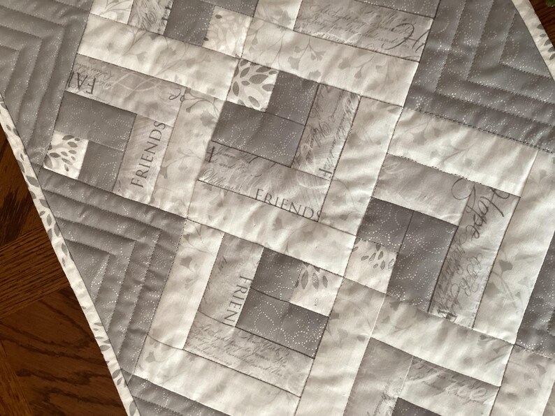 May include: A quilted table runner in shades of gray and white, featuring a geometric pattern. The design incorporates rectangular blocks with the words "Friends" and "Hope" printed on some blocks. The runner is bordered with a silver trim.