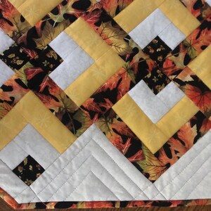 Peut inclure: Chemin de table matelass&eacute; avec un motif r&eacute;p&eacute;t&eacute; de tissu jaune, blanc et imprim&eacute; de feuilles. Le motif pr&eacute;sente un agencement g&eacute;om&eacute;trique de carr&eacute;s et de lignes diagonales, avec une bordure blanche et un bord cousu.