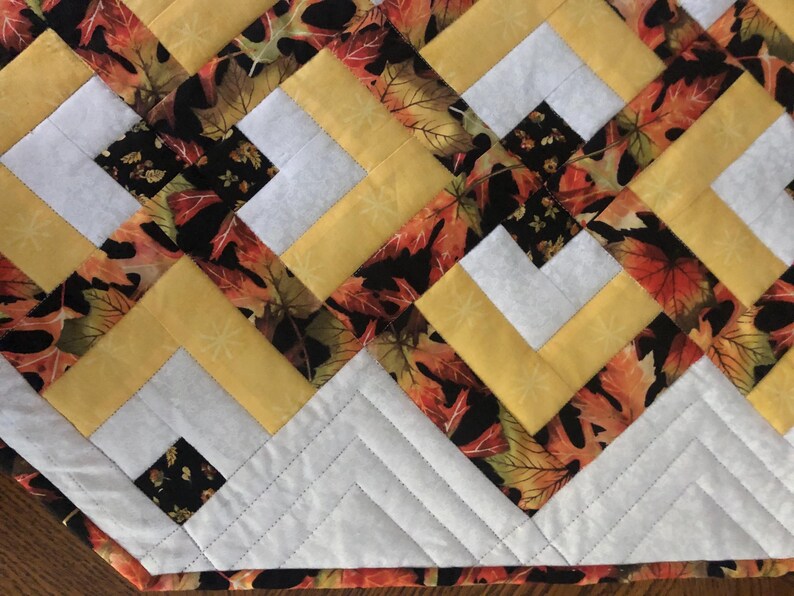 Peut inclure: Chemin de table matelass&eacute; avec un motif r&eacute;p&eacute;t&eacute; de tissu jaune, blanc et imprim&eacute; de feuilles. Le motif pr&eacute;sente un agencement g&eacute;om&eacute;trique de carr&eacute;s et de lignes diagonales, avec une bordure blanche et un bord cousu.