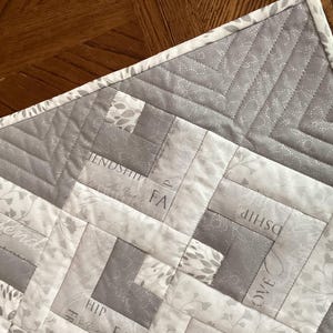 May include: A gray and white quilted fabric with a geometric pattern. The quilt features a log cabin design with the words "Friendship", "Love", and "Faith" subtly printed on the fabric. The border is a light gray with a floral pattern.