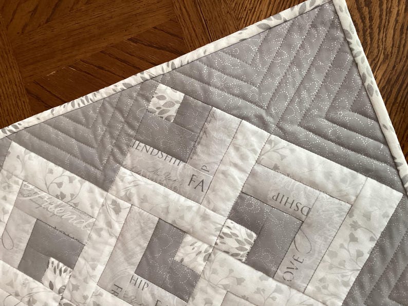 May include: A gray and white quilted fabric with a geometric pattern. The quilt features a log cabin design with the words "Friendship", "Love", and "Faith" subtly printed on the fabric. The border is a light gray with a floral pattern.
