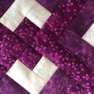 May include: Close-up of a quilted fabric with a woven pattern. The design features alternating squares of white and purple fabric. The purple fabric has a floral leaf pattern. The stitching is white.