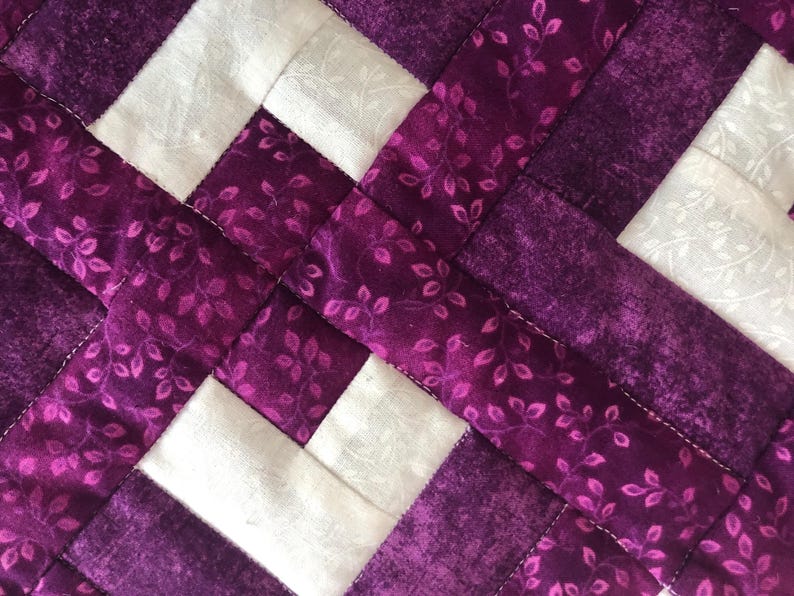 May include: Close-up of a quilted fabric with a woven pattern. The design features alternating squares of white and purple fabric. The purple fabric has a floral leaf pattern. The stitching is white.