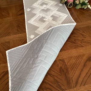 May include: A reversible gray and white quilted table runner with a geometric design. The top side features a pattern of interlocking shapes, while the underside is a solid gray. The edges are finished with a patterned border.