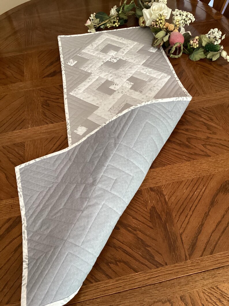 May include: A reversible gray and white quilted table runner with a geometric design. The top side features a pattern of interlocking shapes, while the underside is a solid gray. The edges are finished with a patterned border.