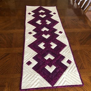 May include: A quilted table runner with a geometric design. The runner features a repeating pattern of white and purple diamond shapes. The purple sections have a speckled texture, and the white sections have diagonal stitching.