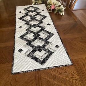 May include: A rectangular quilted table runner featuring a geometric pattern of interlocking black and white squares. The fabric displays a floral print in black on a white background, with intricate chevron-style machine stitching throughout.