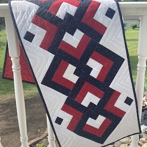 May include: A white table runner with a geometric pattern in red, black, and white. The pattern is made up of squares and rectangles. The runner is hanging on a white railing.