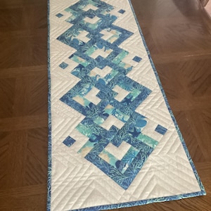 May include: A rectangular table runner featuring a central row of interconnected diamond-shaped quilt blocks in shades of blue, teal, and white, decorated with coral and starfish patterns, bordered by white fabric with a stitched chevron design.