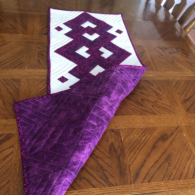 May include: A handmade quilted table runner with a geometric design. The top side features a white background with a purple zig-zag pattern. The underside is a solid purple fabric. The runner is approximately 18 inches wide and 36 inches long.
