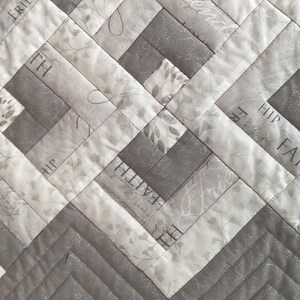 May include: Close-up of a quilted fabric with a geometric design. The quilt features alternating light gray and medium gray rectangular blocks. Some blocks have floral patterns and text that reads "FAITH", "FREE", and "HIP".